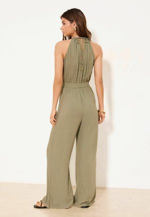 Kobieta stojąca tyłem, ubrana w oliwkowy, bezrękawnikowy kombinezon z odkrytymi ramionami, szerokimi nogawkami i wiązaniem z tyłu.