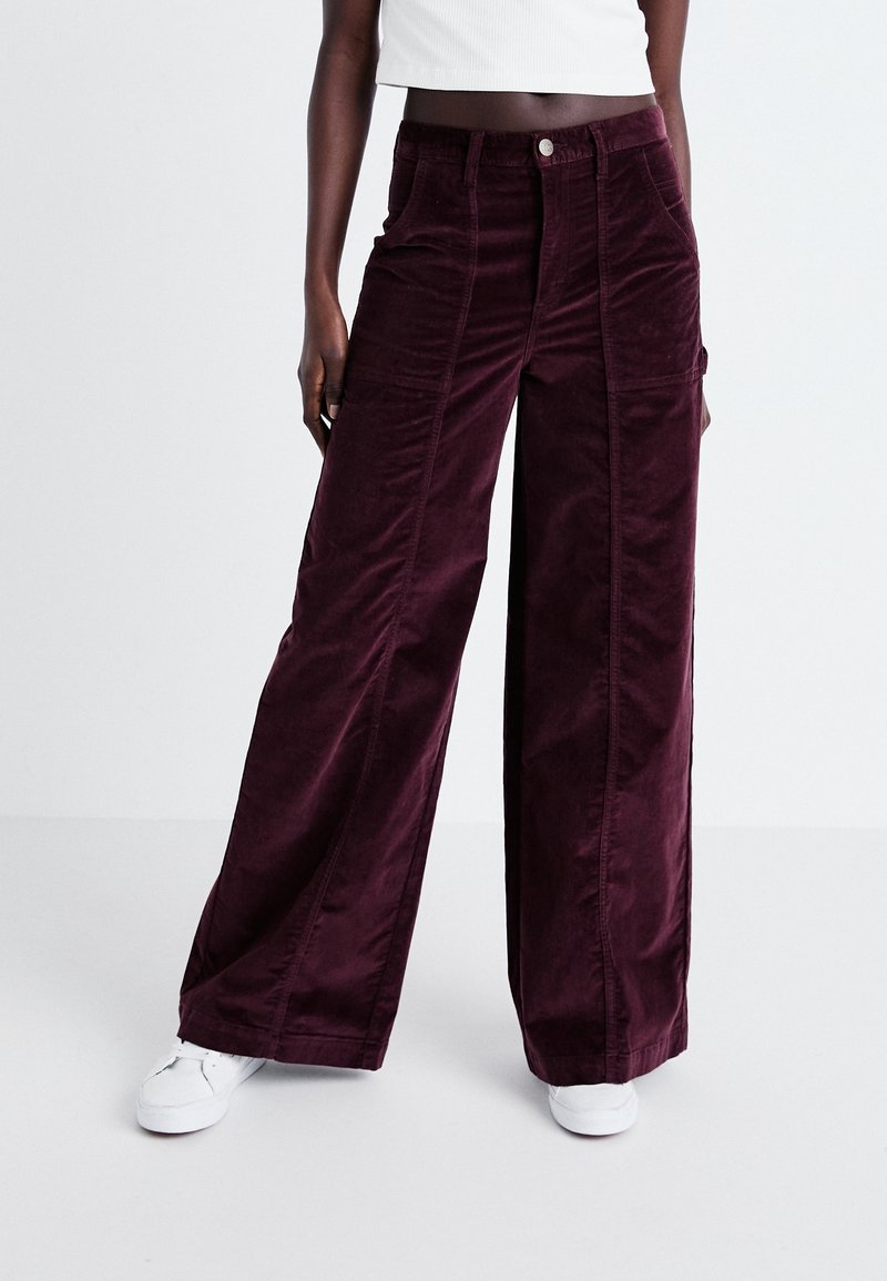 Femme portant un pantalon large en velours côtelé violet foncé et des baskets blanches, debout devant un fond clair uni.