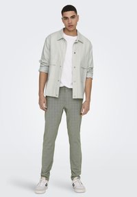 Camisa de botones gris claro, camiseta blanca, pantalones verdes con estampado y zapatillas blancas con detalles en negro. Conjunto casual en general.
