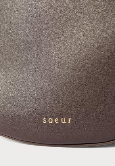 Ruskea nahkakäsilaukku, jossa on sileä pinta ja brändinimi "soeur" kaiverrettuna kultaisena alareunaan. Minimalistinen muotoilu.