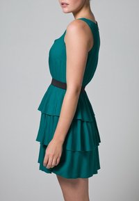 Türkisfarbenes, gestuftes Kleid mit tailliertem Oberteil und ausgestelltem, gerüschtem Rock. Mit einem schwarzen Taillenband und glattem, leichtem Stoff.