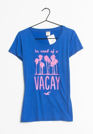 Maglietta di cotone blu a maniche corte, con grafica di una palma rosa e testo "In need of a VACAY." Appesa a un gancio nero.
