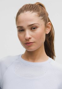 Világoskék sportfelső, amely illeszkedő fazonú, texturált anyagból készült, és hálós részletekkel rendelkezik a vállakon a légáteresztőség érdekében.