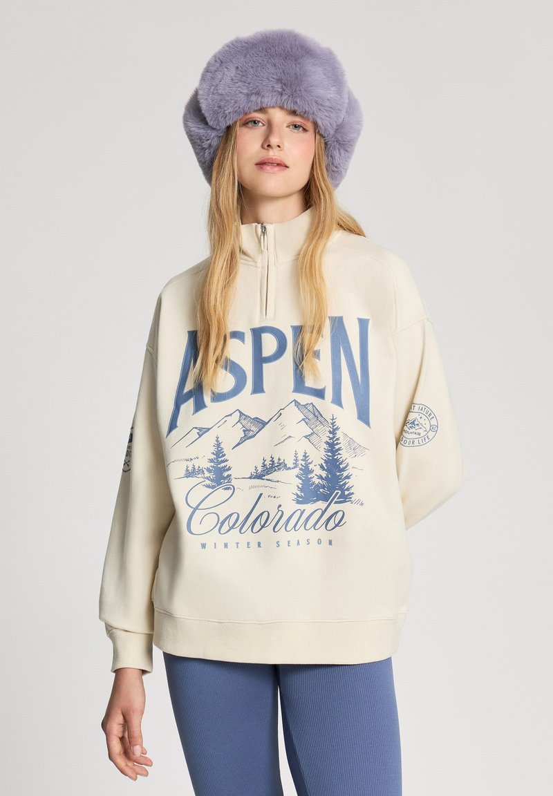 Kremowa bluza z zamkiem na całej długości, z niebieskim napisem i grafiką gór oraz drzew. W komplecie z jasnoniebieskimi prążkowanymi legginsami i fioletową czapką futrzaną.