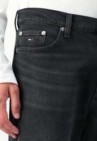 Krupan plan crne traperice s džepom, na kojem se nalazi mali crveni, bijeli i plavi logo, metalni zakovici, a pored njega se nalazi ruka osobe.