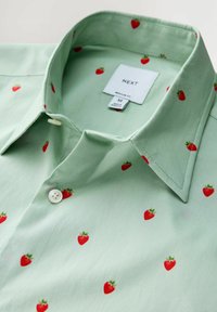 Camisa de color verde claro con un patrón de fresas rojas, abotonada en la parte superior, mostrando la etiqueta de la marca "NEXT" y la etiqueta de tamaño mediana dentro del cuello.