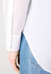 White long-sleeved shirt with a textured fabric, featuring button cuffs. Tucked into light blue denim with a visible back pocket.
