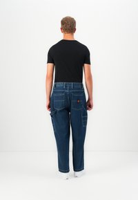 Vīrietis, kurš valkā tumši zilu džinsu stila bikses ar plāksteri uz aizmugures kabatas un melnu īsu piedurkņu kreklu, stāv pretī baltai sienai.