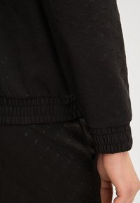 Tessuto nero con texture e un delicato motivo floreale. Presenta polsini ed elastico in vita. Inquadratura ravvicinata della manica e della mano, senza hardware visibile.