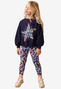 Navy sweatshirt with puffed sleeves featuring a colorful star design. Vibrant, multicolored patterned leggings. Light-colored sneakers.