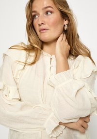 Kreemivärviline pluus volangiliste varrukate ja kaelusega, millel on nööbiplokk ja tekstuuriga kangas. Pehme ja kerge disain.