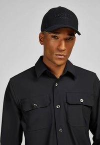 Zwarte knop-up shirt met twee borstzakken, voorzien van zilveren hardware en een soepele textuur, gecombineerd met een donkere geborduurde pet.