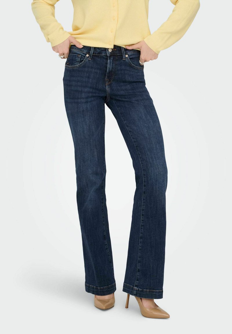 Jean évasé bleu foncé avec cinq poches, en denim, avec des détails tels que des coutures et une ceinture classique. Porté avec un cardigan jaune.