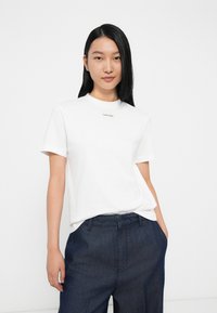 Fiatal nő, aki egyenes fekete hajat visel, fehér Calvin Klein pólóban és sötétkék magas derekú nadrágban, fehér háttér előtt.