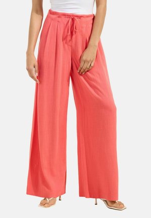 Naine kannab korallikarva laia säärega pükse, millel on nöörvöökoht, ning valgeid kontsakingi, nähtav vöökohast jalgadeni valgel taustal.