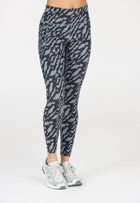Nainen, joka on pukeutunut musta-harmaisiin eläinkuosilla varustettuihin leggings-housuihin ja hopeisiin urheilukenkiin, seisoo valkoista taustaa vasten.