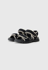 Black sandals with textured fabric straps and cream accents. Thick rubber sole with a rugged tread pattern. Adjustable Velcro closures.