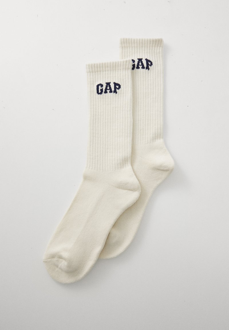 Et par kremfargede ribbestrikkede sokker med "GAP"-logo i mørkeblått nær toppen, på en enkel hvit bakgrunn.