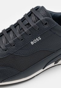 Marinblå lädersneakers med texturerade och släta paneler, runda snörningar och en kontrasterande vit sula. Inkluderar "BOSS"-logotyp på sidan.