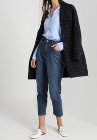 Femme portant un manteau à carreaux noir, un chemisier bleu clair, un jean bleu court taille haute, et des mocassins blancs sans lacets avec un détail argenté.