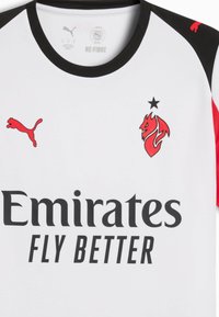 Hvid sportsjersey med sort krave, røde accenter, Puma-logoer og et rødt drageemblem. "Emirates FLY BETTER" trykt med fed sort skrift.