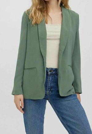 Femme portant un blazer vert clair sur un débardeur côtelé beige, associé à un jean bleu, debout sur un fond clair uni.