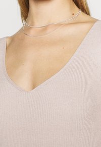 Jasnoróżowa, prążkowana bluzka z dekoltem w kształcie litery V, charakteryzująca się miękką teksturą i dopasowanym krojem. Na tkaninie nałożone są dwa srebrne naszyjniki łańcuszkowe.