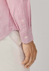 Camisa a rayas rojas y blancas con puños abotonados, que presenta una textura suave y un cuello clásico. Mano descansando sobre pantalones de color claro.