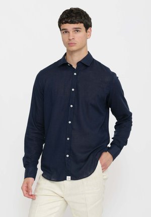 Giovane uomo che indossa una camicia blu navy a maniche lunghe con bottoni e pantaloni color crema, in piedi davanti a uno sfondo bianco semplice.