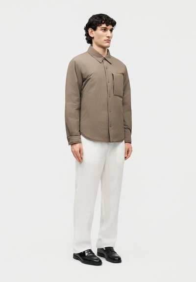 Άνδρας που στέκεται φορώντας καφέ πουκάμισο με κουμπιά και μακριά μανίκια με τσέπη στο μπροστινό μέρος, άσπρο παντελόνι και μαύρα παπούτσια φόρμας σε απλό φόντο.