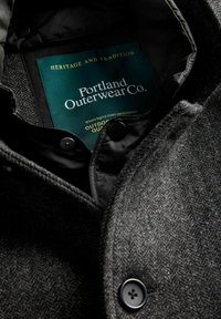 Mørkegrå fiskegrøn uldfrakke med sorte knapper og et grønt Portland Outerwear Co.-mærke inde i kraven.