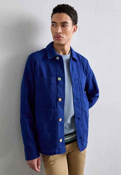Navy blue jacket with a classic collar, two large front pockets, and colorful buttons. Worn over a light blue sweater, paired with beige pants.