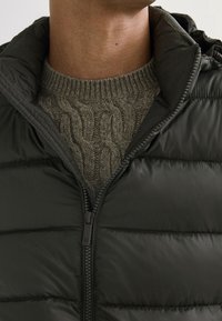 Dunkelgrüne Steppjacke mit horizontalem Steppmuster und hohem Kragen, offen getragen, sodass ein hellgrauer Strickpullover darunter sichtbar ist.