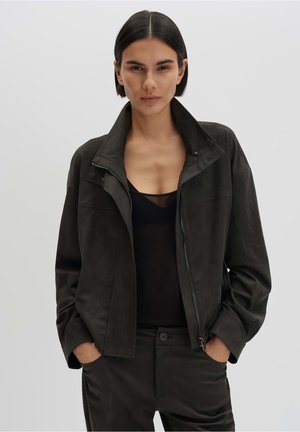 Frau mit kurzen dunklen Haaren, die eine offen getragene dunkle Jacke, ein schwarzes Top und passende dunkle Hose trägt, mit den Händen in den Taschen vor einem einfarbigen Hintergrund.