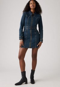 Abito in denim con maniche lunghe, cerniera frontale, vita aderente e due tasche laterali. Colore blu scuro con cuciture a contrasto. Abbinato a stivali neri alla caviglia.
