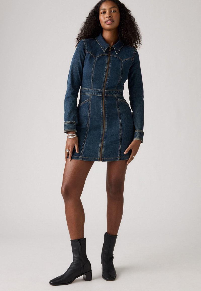 Abito in denim con maniche lunghe, cerniera frontale, vita aderente e due tasche laterali. Colore blu scuro con cuciture a contrasto. Abbinato a stivali neri alla caviglia.