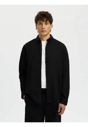 Mann med krøllete hår iført en svart knappeskjorte over en hvit t-skjorte, stående mot en enkel lys bakgrunn.