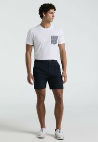 Weißes T-Shirt mit einer gemusterten Tasche, kombiniert mit marineblauen Shorts. Model trägt weiße Sneaker und steht vor einem schlichten Hintergrund.