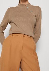 Beige Langarmpullover mit hochgeschlossenem Kragen und diagonaler Knopfdetaillierung; kombiniert mit tanfarbenen, hochgeschnittenen Hosen mit einer glatten Textur.