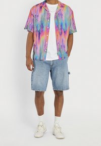 Camisa de manga corta colorida con patrones ondulados en rosa, azul y verde; combinada con pantalones cortos de mezclilla azul claro y zapatillas blancas.