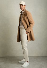 Man wearing beige long coat, cream buttoned sweater, off-white pants, white sneakers, and white cap, standing against neutral background.
