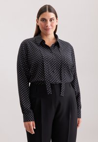 Zwarte overhemd met witte stippen, met lange mouwen en een kraag. Gecombineerd met zwarte hoge taille broeken.