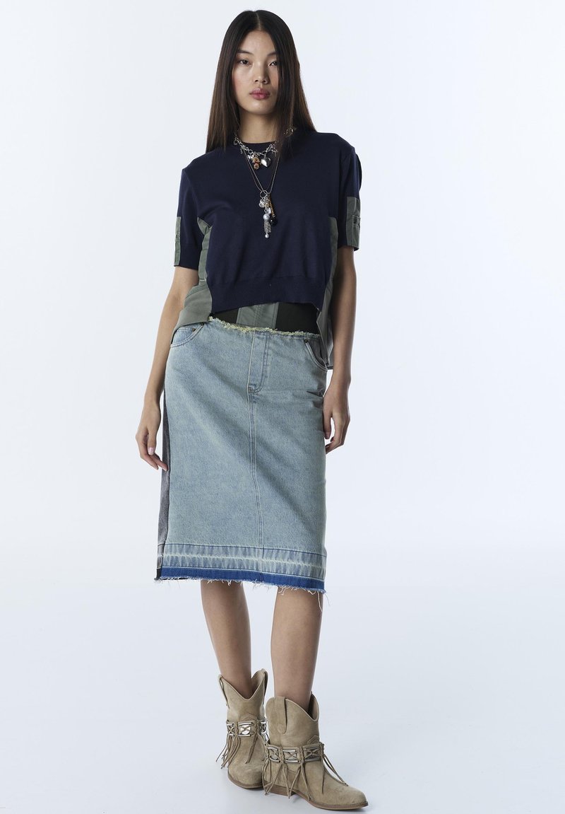 Mujer joven con una blusa de manga corta azul marino y oliva, falda midi de mezclilla, collares en capas y botines de ante beige con flecos.