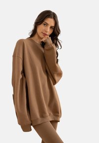 Brązowy oversize'owy sweter wykonany z miękkiego materiału, z okrągłym dekoltem, długimi rękawami i ściągaczami. Model pozujący z ręką na brodzie.