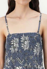 Blaues Blumenkleid mit weißen Mustern, gerafftem Oberteil, dünnen Trägern und strukturiertem Stoff. Akzente von Punkten an den Kanten der Träger.