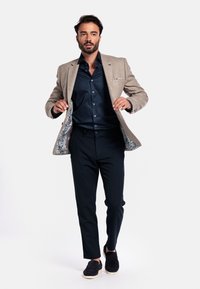 Beige blazer with light gray lining, blue shirt, dark trousers, and black slip-on shoes. Blazer has a subtle texture and embroidered detail.