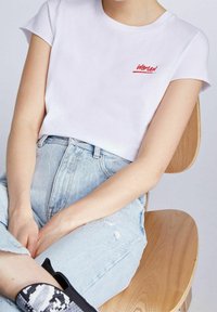 Camiseta de algodón blanca de manga corta con texto rojo que dice "MUJER" en la zona del pecho izquierdo, combinada con unos vaqueros de mezclilla azul claro.