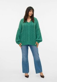 Pull en tricot vert avec un col en V, orné de motifs en cables texturés, associé à un jean bleu clair évasé et des chaussures brunes à bouts pointus.