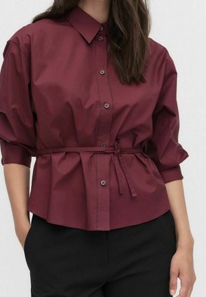 Femme portant une chemise bordeaux à boutons avec manches retroussées et une ceinture nouée à la taille, associée à un pantalon noir.