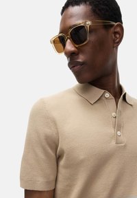 Camisa de polo de punto beige con mangas cortas y tapeta de tres botones; usando gafas de sol de montura transparente con lentes marrones.